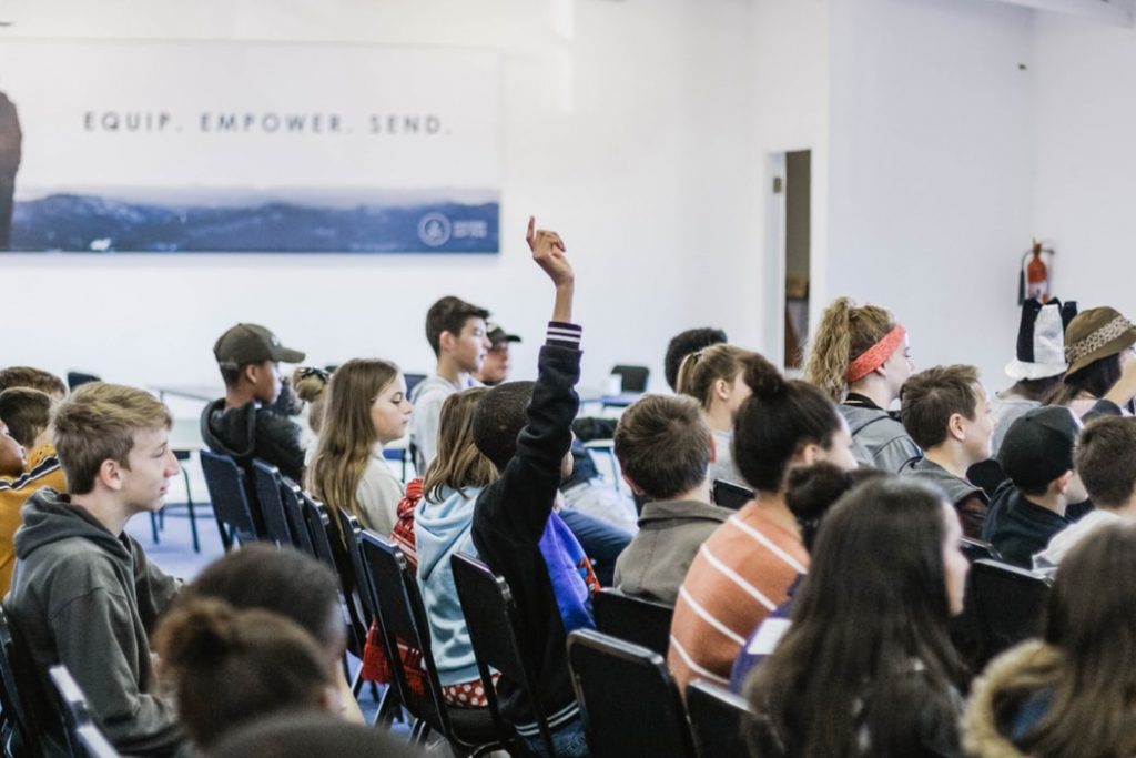 A room full of about twenty teenage students with one in the centre raising their hand